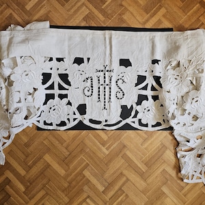 May include: An off-white linen altar cloth featuring intricate cutwork embroidery of floral patterns and the central monogram IHS with a cross. The fabric has a wide, solid top border and decorative scalloped edges with detailed needlework.