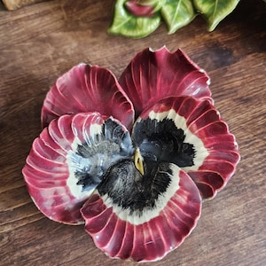 Op de afbeelding: Een keramisch viooltje-ornament met dieprode bloemblaadjes en een zwart-wit patroon in het midden. De bloem heeft een glanzende afwerking en een sculpturaal, gelaagd ontwerp dat zorgt voor een realistische, driedimensionale uitstraling.