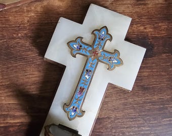 May include: A white marble cross with a decorative blue, gold, and red cross overlay. The cross has a small, ornate, gold-trimmed basin at the base. An open book rests in the background on a wooden surface.