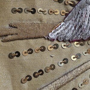 May include: Close-up of a gold fabric with intricate embellishments. The fabric is adorned with rows of small, dark-colored sequins and gold thread detailing. A section of silver sequins and a gold thread design are visible.