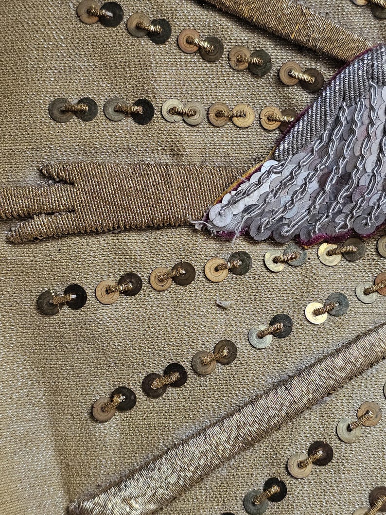 May include: Close-up of a gold fabric with intricate embellishments. The fabric is adorned with rows of small, dark-colored sequins and gold thread detailing. A section of silver sequins and a gold thread design are visible.