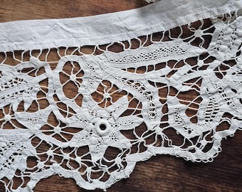 May include: Close-up of a white lace trim with intricate floral and geometric patterns. The lace features a scalloped edge and is attached to a plain fabric border. The delicate design is set against a dark wood surface.