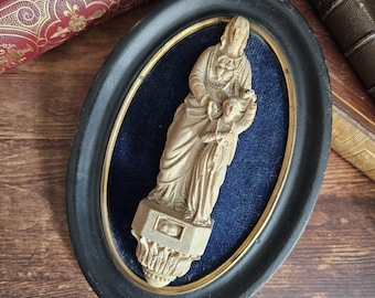May include: An antique oval-framed relief sculpture featuring a carved figure holding a child, set against a blue velvet background. The frame is black with a gold inner edge. The sculpture is a light beige colour.