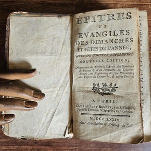 May include: An antique book titled "ÉPITRES ET ÉVANGILES DES DIMANCHES ET FÉTES DE L'ANNÉE, AVEC DE COURTES RÉFLEXIONS, NOUVELLE ÉDITION" with a decorative floral design on the cover. The book was published in Paris in 1774.
