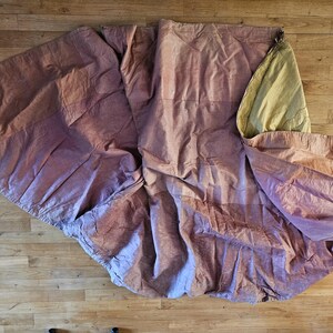 May include: A large, draped fabric in shades of mauve and tan lies on a wooden floor. The fabric is bunched and folded, with visible seams and a lighter-colored section at the top. The texture appears soft and possibly linen or cotton.