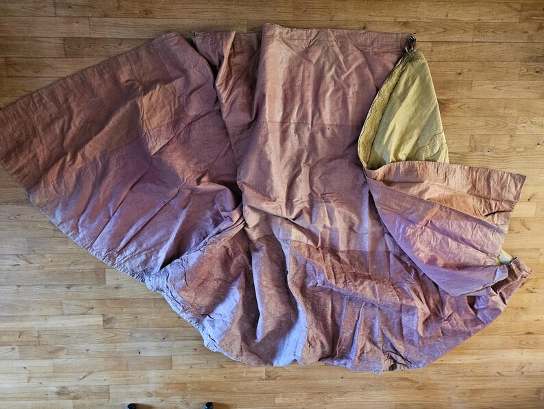 May include: A large, draped fabric in shades of mauve and tan lies on a wooden floor. The fabric is bunched and folded, with visible seams and a lighter-colored section at the top. The texture appears soft and possibly linen or cotton.