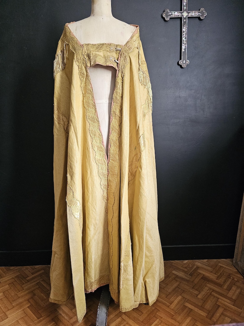 May include: A long, flowing, golden-yellow cape with intricate gold embroidery and fringe detailing. The cape is draped over a mannequin, showcasing its design and texture. The garment has a pink lining and is likely vintage or antique.