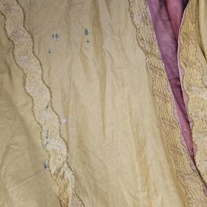 May include: Close-up of a vintage garment with a pale yellow exterior and a deep pink interior. The yellow fabric has a subtle sheen and is trimmed with delicate lace. There are a few small blue stains on the yellow fabric.