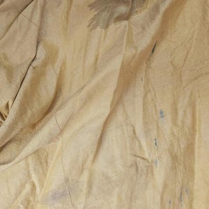 May include: Close-up of a tan fabric with visible folds and creases. The fabric has dark stains and blue marks scattered across its surface. The texture appears smooth, with a subtle sheen. The edges of the fabric are not visible.