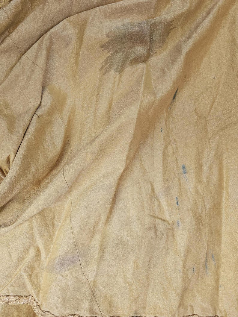 May include: Close-up of a tan fabric with visible folds and creases. The fabric has dark stains and blue marks scattered across its surface. The texture appears smooth, with a subtle sheen. The edges of the fabric are not visible.