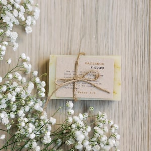 Op de afbeelding: Een handgemaakt stuk zeep met een papieren label vastgebonden met touw. Het label leest "PATIENCE" in het Engels en Hebreeuws, met een bijbelvers. Witte gipskruidbloemen zijn rond de zeep gerangschikt op een houten oppervlak.