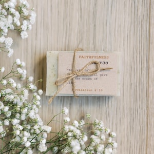 May include: A stack of handmade soap bars tied with twine, featuring a small card with the word "FAITHFULNESS" and a Bible verse. The soap is light green with visible herbs. White flowers are arranged around the soap on a wooden surface.