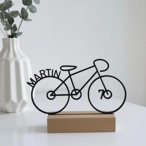 May include: Black metal bicycle sculpture with the name "MARTIN" and the number "70" integrated into the design. The sculpture is mounted on a rectangular wooden base. A white vase with greenery is in the background.