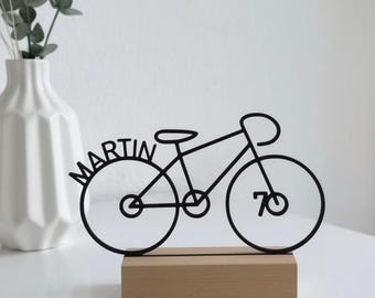 Könnte beinhalten: Schwarze Metall-Fahrradskulptur mit dem Namen "MARTIN" und der Zahl "70" im Design integriert. Die Skulptur ist auf einem rechteckigen Holzsockel montiert. Im Hintergrund eine weiße Vase mit Grün.