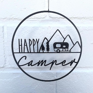 May include: A circular black metal wall hanging featuring a silhouette design of mountains, two pine trees, and a travel trailer. The text "HAPPY" is printed in a sans-serif font above the trailer, and the word "Camper" is in cursive below.