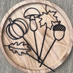 Könnte beinhalten: Ein rundes Holztablett mit vier schwarzen, drahtartigen Silhouetten von herbstlichen Elementen: ein Kürbis, ein Pilz, ein Ahornblatt und eine Eichel. Das Tablett hat eine natürliche helle Holzmaserung und einen erhöhten, kreisförmigen Rand.