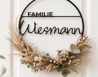 Könnte beinhalten: Ein schwarzer, runder Metallring mit dem Schriftzug FAMILIE und Wiesmann in Schreibschrift. Die untere Hälfte des Rings ist mit getrockneten beigen Gräsern, kleinen weißen Blüten, runden Samenständen und grünen Eukalyptusblättern verziert.