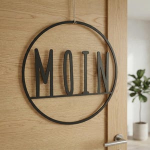 May include: A black circular wall hanging featuring the word MOIN in capital letters. The sign is constructed from a thin metal frame and is suspended by a light-colored string. The letters are integrated into the circular frame design.