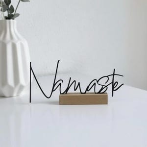 May include: A black metal sign that reads "Namaste" in cursive script, mounted on a small, rectangular wooden base. A white vase with a geometric design and greenery is in the background.