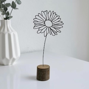 May include: A black wire daisy sculpture with a round flower head and a long stem, mounted on a small, cylindrical wooden base. The sculpture is a minimalist, modern home decor item.