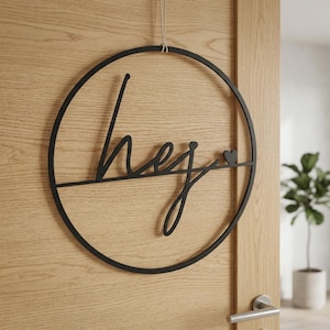 Könnte beinhalten: Ein schwarzer, kreisförmiger Wandbehang mit dem Wort hej in einer kursiven Schriftart. Das Design umfasst eine horizontale Linie, die durch die Mitte des Kreises verläuft, und ein kleines Herzsymbol rechts neben dem Text, aufgehängt an einer Schnur.