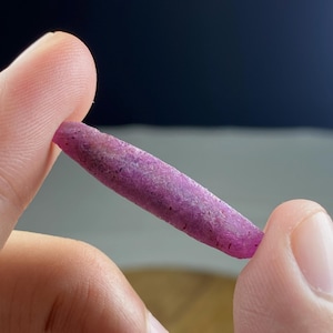 Puede incluir: Una gema en bruto, alargada, de color rosa púrpura, se sostiene entre dos dedos. La piedra tiene una superficie rugosa y texturizada con inclusiones más oscuras. El fondo es un degradado borroso de azul oscuro y gris claro.