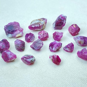 May include: A collection of raw ruby gemstones in varying sizes and shapes. The rubies display a deep pink to red hue with some white and grey inclusions. The gemstones are scattered on a light-colored surface.