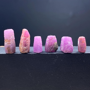 May include: Six rough ruby gemstones in varying sizes and shapes. The gems display shades of pink and red, with some showing a crystalline structure and others appearing more opaque. They are arranged on a dark surface, showcasing their natural beauty.
