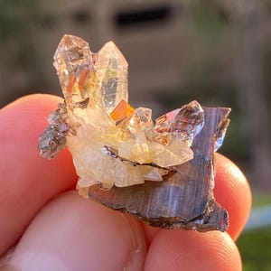 Puede incluir: Un grupo de cristales naturales, con formaciones de cuarzo claras y puntiagudas con toques de naranja y marrón. Los cristales se asientan sobre un fondo de depósitos minerales más pequeños y de color claro y una base oscura y metálica. La imagen muestra la belleza natural de los especímenes minerales.
