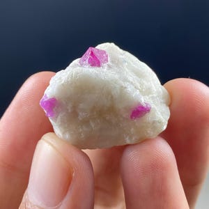 May include: A raw, uncut gemstone featuring a cluster of vibrant pink ruby crystals embedded in a white matrix. The rough stone displays a natural, unpolished texture, with the ruby crystals showcasing a brilliant, crystalline structure.