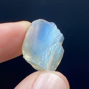 May include: A translucent, rough-cut gemstone with a pale blue and white color gradient. The stone has a natural, irregular shape and is held between two fingers against a dark background. The gemstone appears to be a raw, uncut mineral.