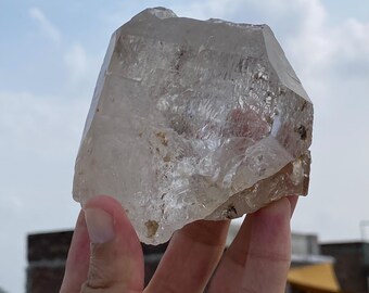Puede incluir: Un gran cristal de cuarzo transparente con una superficie rugosa y texturizada. El cristal se sostiene en una mano contra un fondo de cielo nublado. Las facetas del cristal capturan la luz, mostrando su forma natural.