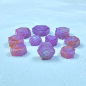 Op de afbeelding: Een verzameling van negen roze en witte gefacetteerde edelstenen, elk met een zeshoekige vorm. De edelstenen zijn in een cirkel op een wit oppervlak gerangschikt.