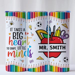 May include: A white tumbler with a colorful pencil border and the text "IT TAKES A BIG HEART TO SHAPE LITTLE MINDS." Features an apple with school supplies and the name "MR. SMITH."