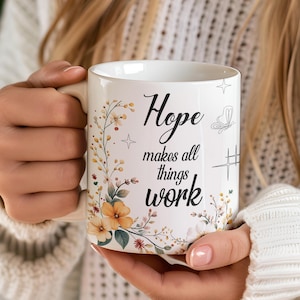 May include: White ceramic mug with the words "Hope makes all things work" in black script. The mug features floral and butterfly illustrations in soft colors. A star and a hashtag-like symbol are also present.