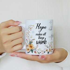 May include: White ceramic mug with the text "Hope makes all things work" in black script. The mug is decorated with floral and botanical illustrations in shades of yellow, orange, and green. The mug is held in both hands.