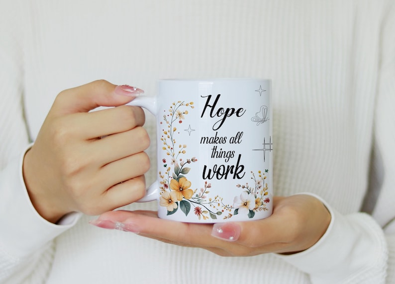 May include: White ceramic mug with the text "Hope makes all things work" in black script. The mug is decorated with floral and botanical illustrations in shades of yellow, orange, and green. The mug is held in both hands.