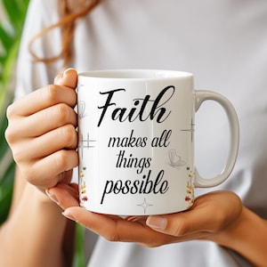 May include: White ceramic mug with the words "Faith makes all things possible" in black script. The mug has a handle and is decorated with small floral and butterfly designs. The mug is held by a person.