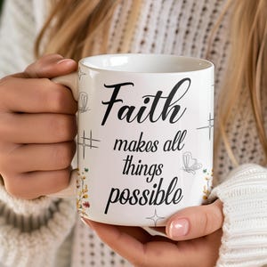 May include: White ceramic mug with the words "Faith makes all things possible" in black script. The mug has decorative floral and butterfly accents. The mug is held by a person wearing a white sweater.