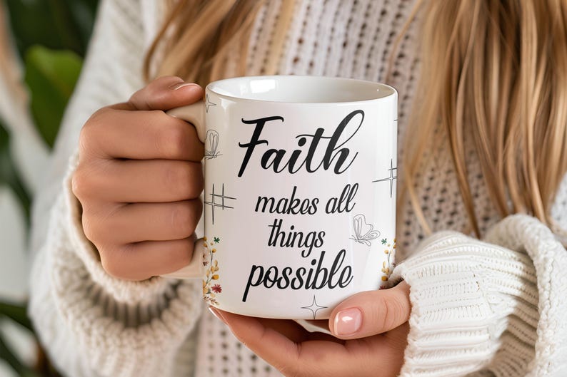 May include: White ceramic mug with the words "Faith makes all things possible" in black script. The mug has decorative floral and butterfly accents. The mug is held by a person wearing a white sweater.