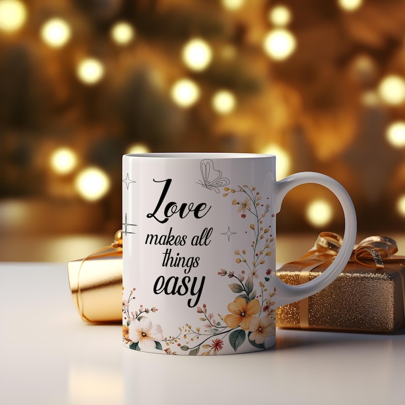 May include: White ceramic mug with the text "Love makes all things easy" in black script. The mug is decorated with floral and butterfly designs in yellow and green. A gold gift box is in the background.