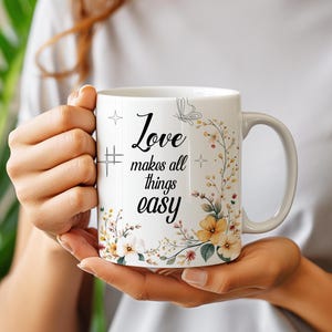 May include: White ceramic mug with the text "Love makes all things easy" in black script. The mug features a floral design with yellow and white flowers and green leaves. A butterfly and star accents are also included.