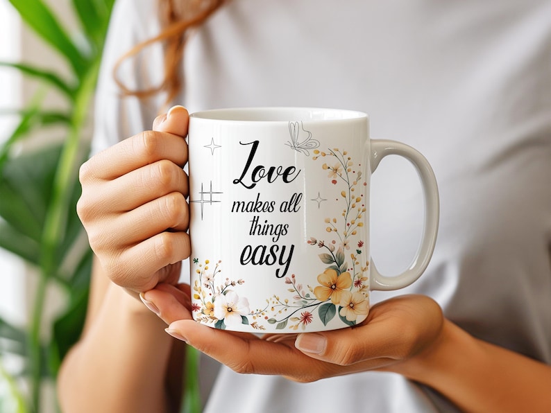 May include: White ceramic mug with the text "Love makes all things easy" in black script. The mug features a floral design with yellow and white flowers and green leaves. A butterfly and star accents are also included.