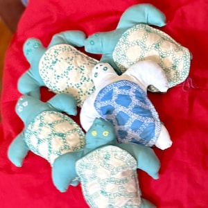 Peut inclure: Cinq jouets en peluche en forme de tortue faits à la main dans différentes nuances de bleu et de vert avec des motifs brodés blancs. Les tortues sont posées sur une surface en tissu rouge.
