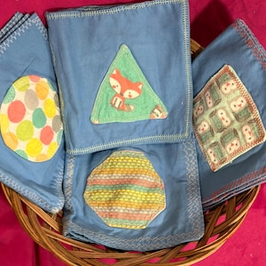 Könnte beinhalten: Blaue Stoffservietten mit bunten Applikationen, darunter ein Fuchs, Punkte und geometrische Muster. Die Servietten sind in einem Weidenkorb angeordnet und zeigen einen handgefertigten Stil.
