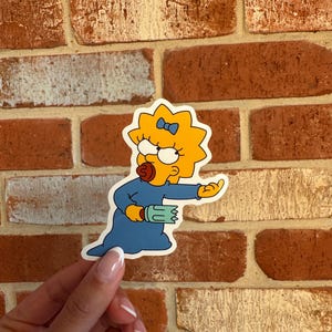 May include: A die-cut sticker of Lisa Simpson from The Simpsons. The character is shown with yellow skin, wearing a blue dress and a blue bow. She is holding a green object and pointing. The sticker is set against a red brick wall.