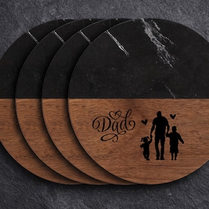 May include: A set of four round coasters featuring a split design with black marble on the top half and brown wood on the bottom half. The wood section is engraved with the word Dad in script, two small hearts, and a silhouette of an adult holding hands with two children.