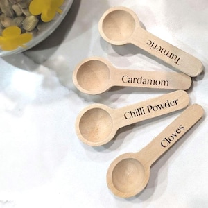May include: Four small wooden spice spoons with text labels: Turmeric, Cardamom, Chilli Powder, and Cloves. The spoons are light brown and arranged on a white surface. A glass jar with yellow flower decorations is visible in the background.
