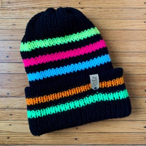May include: Black knit beanie with colorful stripes in neon green, pink, blue, and orange. The beanie has a small tag that says "Hug Hats" with a heart.