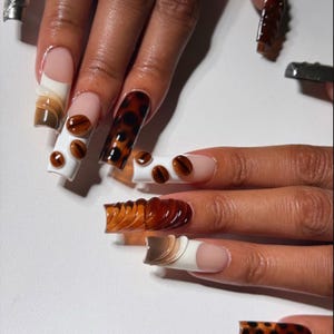 May include: A set of long, sculpted acrylic nails with various designs. Some nails feature a coffee bean design, others have a tortoiseshell pattern, and some have a layered, textured appearance in shades of brown and white. The nails are on a light background.